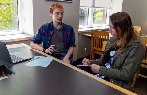 A writing consultant with short neon-colored hair explains something to another student who's writing in a notebook.