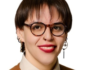 Monica Cornejo smiles at the camera. She is wearing glasses, a collared shirt, and several earrings. 