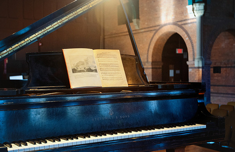 A black grand piano on a wooden stage in a large hall with brick archways and green trim. The piano lid is open, and a sheet of music rests on the music stand. Warm stage lighting illuminates the piano, creating a dramatic contrast with the darker seating area in the background.