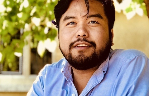 Close up of a person wearing a light blue button down shirt smiling into the camera. Natural light and foliage can be seen in the background.