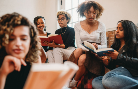 A diverse group of five young women sit closely together, engaged in reading books. They appear focused and relaxed in a warmly lit room by a window.