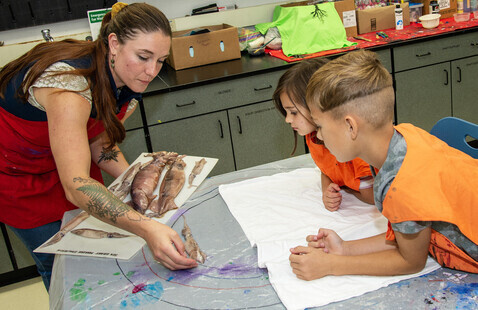Two children look on as an instructor shows them how to paint while holding a squid.