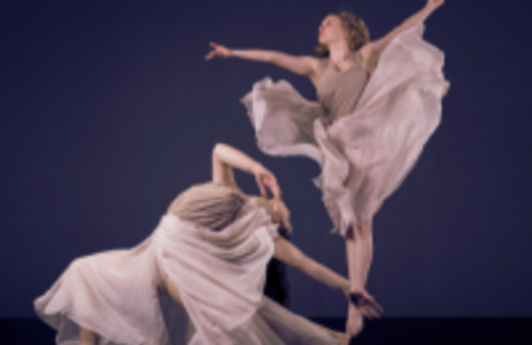Two dancers dressed in tan dresses. One jumping and the other lunged out with head back.