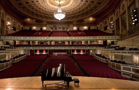 piano on stage with seating and large chandelier in background.