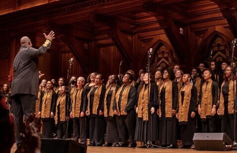 Kuumba Singers on stage at Sanders Theatre with conductor Sheldon Reid '97 in front with his arm raised.