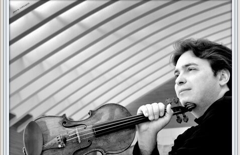 Black-and-white photograph of a musician holding a violin at shoulder height, with curved architectural lines sweeping across the background and the instrument prominently framed in the foreground.