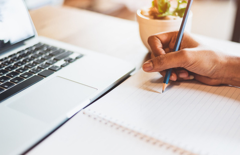 Shutterstock image of someone writing in a notebook with a laptop in view.