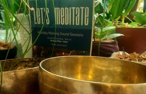 2 hammered brass Tibetan singing bowls in the foreground. A paper sign with the words "Let's Meditate" and  several tropical plants in the background.