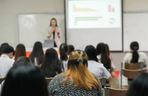 Group of people at a presentation sitting in desks, person in front presenting with a pull-down screen