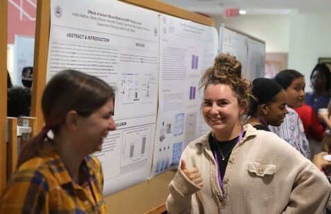 photo of a group of students standing in front of a research poster, one is smiling and giving a thumbs up at the camera