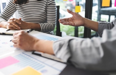 Two people converse at a table covered with documents and colorful sticky notes, suggesting a collaborative and focused discussion in a bright office.