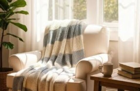 A softly lit interior scene featuring a light-colored upholstered armchair placed beside a large window. Sunlight streams through sheer white curtains, illuminating the chair and wooden floor. A striped knit blanket in neutral gray and cream tones is draped over the chair. To the right of the chair is a small wooden side table holding a ceramic mug and several stacked books. A leafy green plant stands nearby. The overall setting appears calm, warm, and quiet, with natural daylight filling the space.
