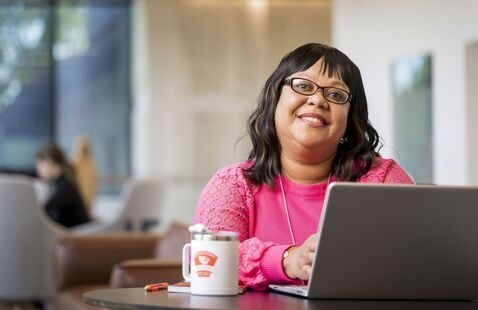Smiling woman sitting at a desk with a laptop and mug of coffee in front of her. 