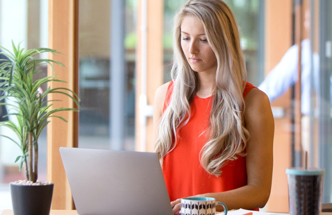 Woman in red shirt sitting at a table with a laptop and mug in front of her. She is typing on the laptop.