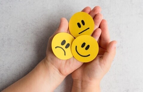 Two hands holding three round yellow emojis, happy, sad and disappointed.