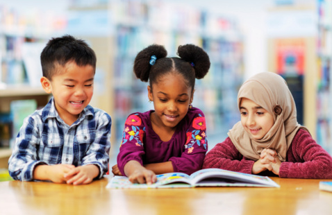 Three smiling children from different cultural backgrounds sit at a table in a library, looking at a picture book together. They appear engaged and excited while learning and sharing the reading experience.