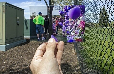 The image shows a hand holding a smaller photograph up against a larger scene. The background features a chain-link fence on the right and a green electrical box on the left, with a small area of brown mulch in between. The smaller photograph, held between the thumb and fingers, portrays a vibrant scene with people and numerous purple balloons and flowers.