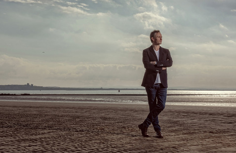 A person wearing a dark suit standing on a beach with a body of water and the sky in the background.
