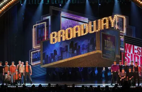 A set broadway stage. The set resembles a theater, flanked with lights and cast members.