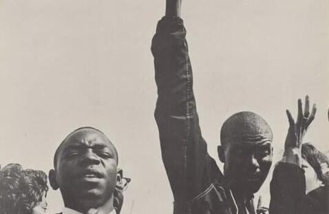 A black and white photo of two young Black men, one with a raised arm, and the word “NOW” at the top.