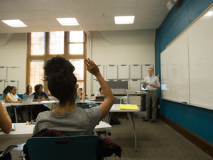 A student raises their hand in class, a professor in the backgroud, Mount Holyoke.