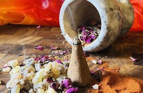Wisps of smoke rise from a cone-shaped incense, which is surrounded by frankincense resin, dried purple petals, and powdered spices laid out on a wooden surface. Behind the incense is a tipped marble mortar and a vivid orange-red fabric.