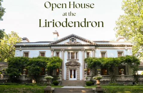 A white and tan mansion with pillars stands surrounded by greenery and trees