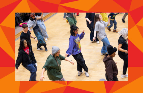 A wood-floor dance studio filled with dancers in sweatshirts, jeans, and sneakers.