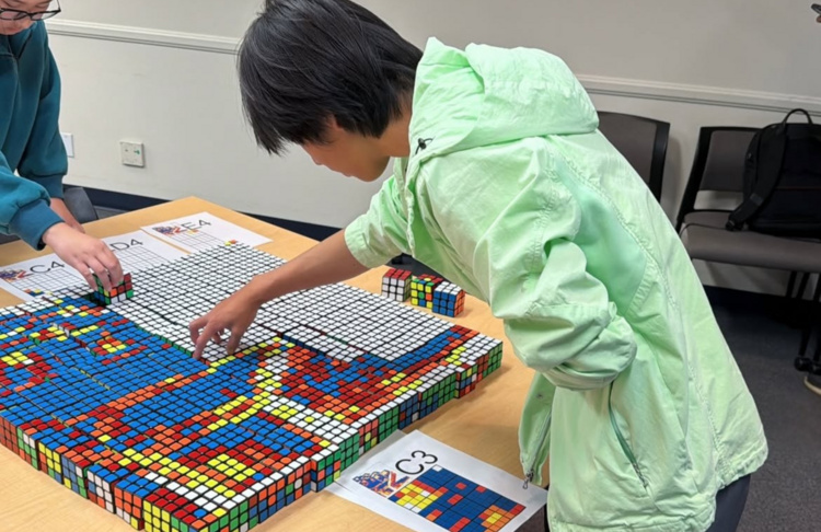 Grid of Rubik's Cubes laid out on a table with two people placing cubes into the gird. 