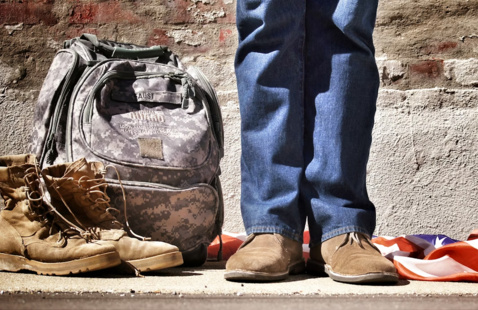 photo of person in jeans shown from knees down, next to military backpack and soldier's boots. There is an American flag fallen on the ground behind the person. 