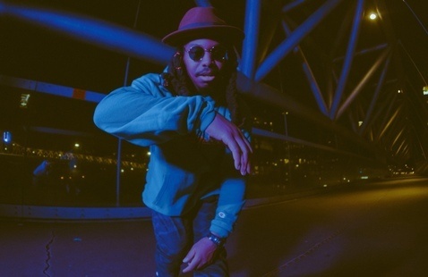 Stiggity Stackz in blue tones bending towards the camera with sunglasses and a hat.