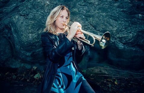 A woman wearing a black leather jacket and patterned blue dress plays a trumpet outdoors, standing in front of a rocky stone background and focused intently on the instrument.