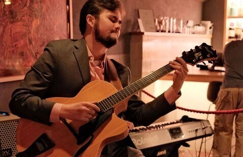 A musician seated indoors plays an acoustic guitar during an intimate concert, with a keyboard, amplifier, and softly lit bar area visible in the background.