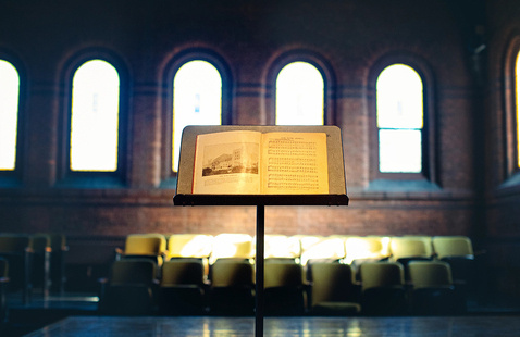An open music score resting on a music stand at center stage, with rows of empty chairs behind it and tall, arched windows letting in soft light inside a brick-walled performance hall.
