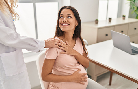 Pregnant woman sitting in a medical office smiles up at a healthcare provider who gently rests a hand on her shoulder, with a laptop on the desk nearby.