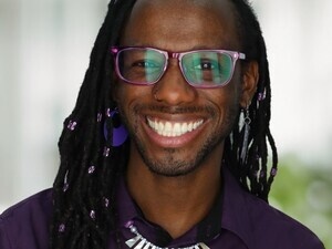 Nick looks at the camera smiling. He is wearing a deep purple button down shirt, a silver necklace, purple earrings, and purple framed glasses.