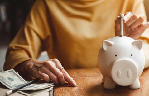 person in a yellow top putting cash into a white piggy bank on the table with a notebook, some other cash, and a pencil also on the table