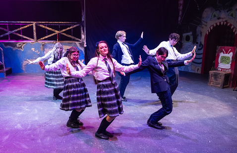 Six theatre performers on stage in school uniforms posing energetically under purple lights with a funhouse themed background.