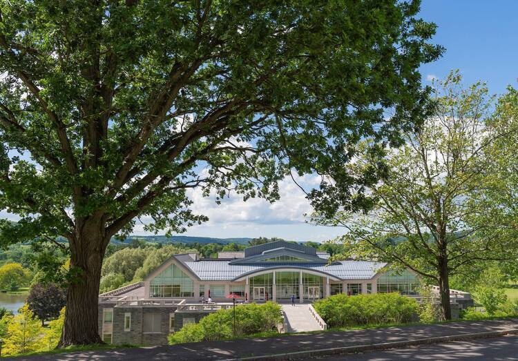 A large, modern stone and glass building with a curved metal roof is nestled into a lush green hillside under a bright blue sky. Massive leafy trees in the foreground frame the view, which overlooks a peaceful valley with a distant lake and rolling hills.