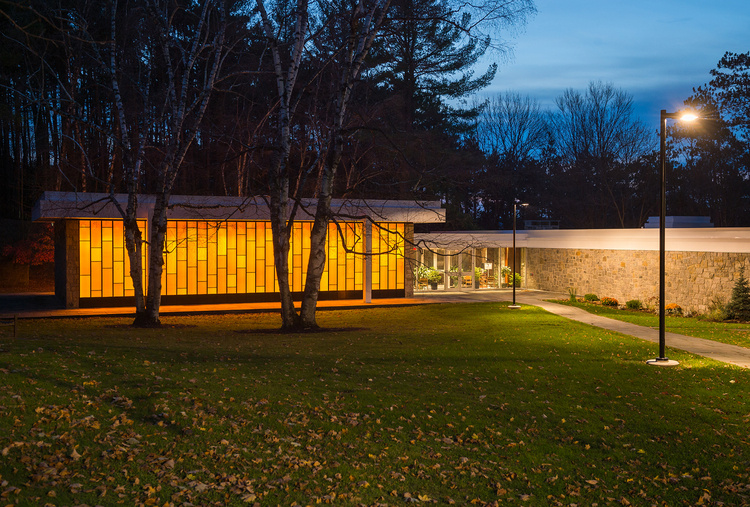 A modern stone building with a flat roof is captured at dusk, featuring a large wall of glowing orange, vertical rectangular windows that cast a warm light over the lawn and birch trees. A stone walkway illuminated by a street lamp leads toward a glass-walled entrance, all set against a backdrop of dark, silhouetted pines and a deep blue evening sky.