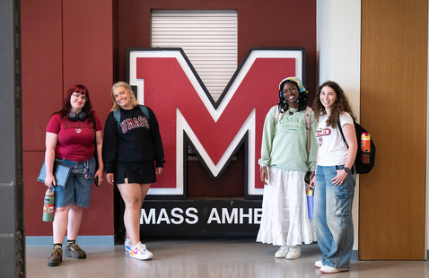 four students pose in front of a large standup sign featuring the UMass Amherst collegiate M logo