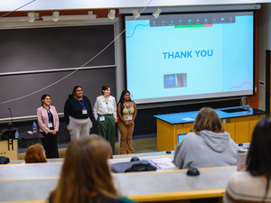 Panel of presenters stand together, presentation in the background, audience in foreground.