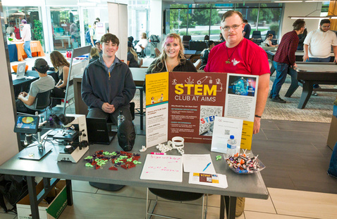 Three individuals standing behind a table with a tri-fold poster reading "STEM Club at Aims", and various other papers and science equipment on the table