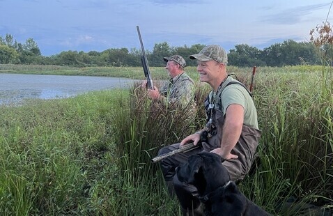 Two duck hunters smiling while they sit in grass next to water