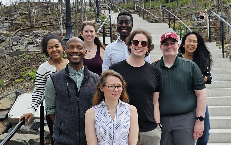 A diverse group of eight smiling individuals poses for a group photo while standing on a wide, concrete outdoor staircase. The group is arranged in layers against a background of a terraced, rocky hillside with budding springtime vegetation and several black lampposts.