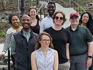 A diverse group of eight smiling individuals poses for a group photo while standing on a wide, concrete outdoor staircase. The group is arranged in layers against a background of a terraced, rocky hillside with budding springtime vegetation and several black lampposts.