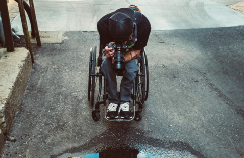 Wheelchair user photographing a puddle that reflects their silhouette, a building, and the sky.