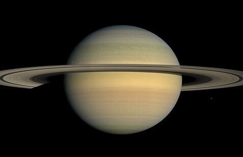 The planet Saturn, a yellow orb, encircled by rings, floating against the black void of space.