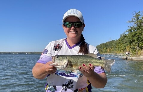 A woman in a hat and sunglasses holding a bass and smiling at the camera.