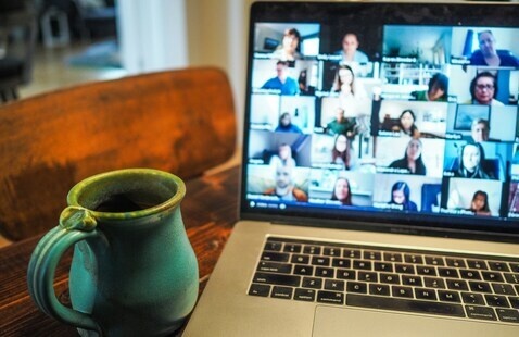 A coffee cups sits beside a laptop. On the  laptop's screen is people participating a webinar.